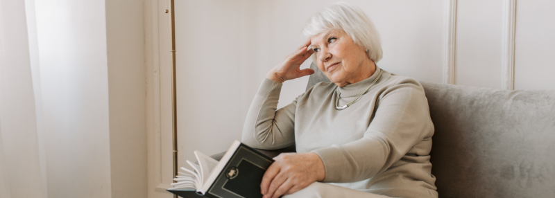 Elderly woman sitting on the couch with a book in her hand, looking concerned