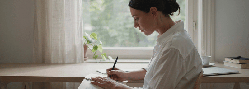 Woman writing in diary by the desk with laptop and cup in back